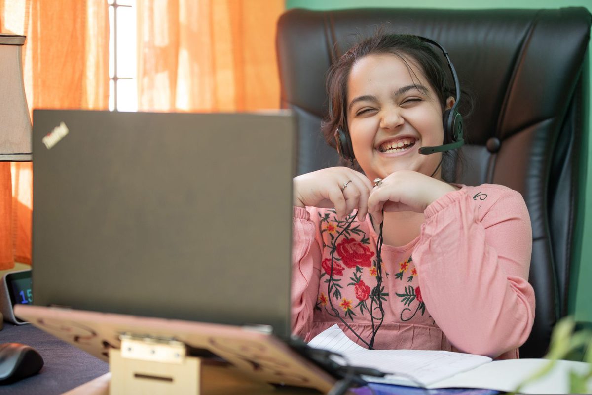 A schoolgirl in uniform attending a live class on a phone propped against her textbooks at home